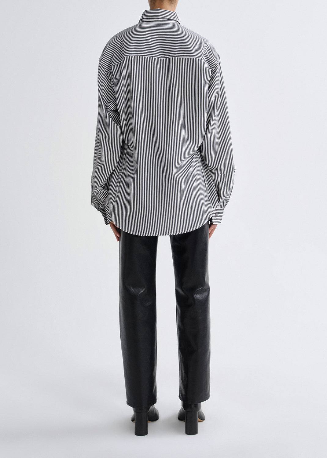 A model shown from the back wearing a black and white vertically striped button-down shirt and black leather trousers, photographed against a grey background.