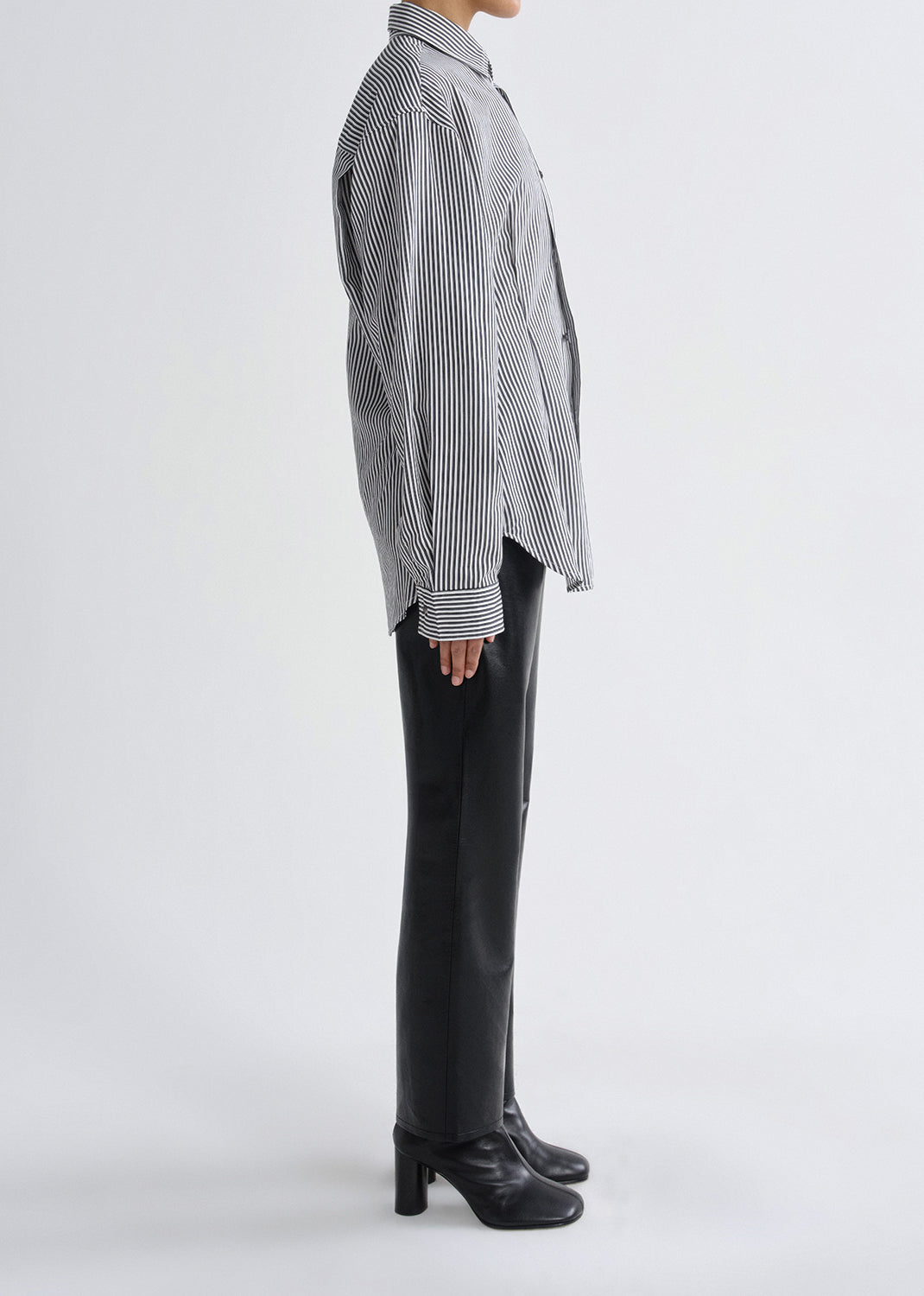 A model shown from the side wearing a black and white vertically striped button-down shirt, black trousers, and black heeled ankle boots against a grey background.