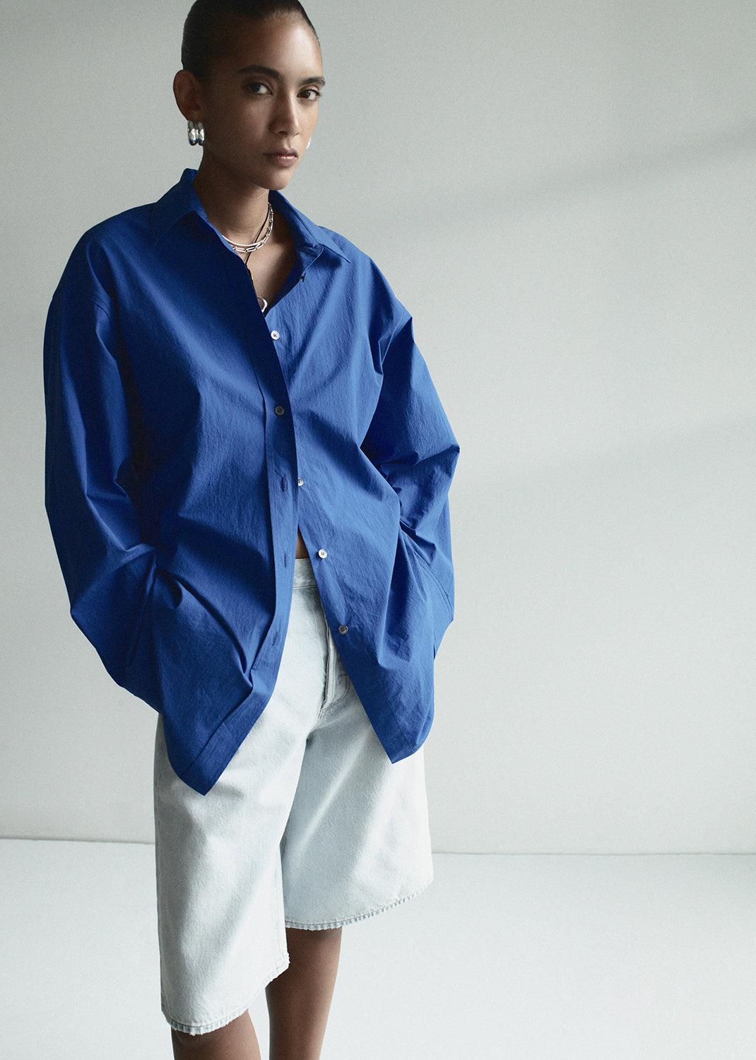 Person wearing a blue oversized shirt and white shorts on a plain background