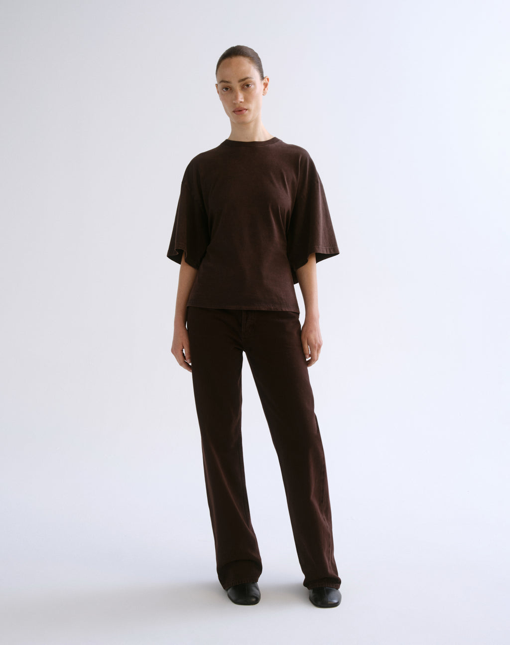 A model stands against a light gray studio background wearing a matching dark brown short-sleeve top and wide-leg trousers set.