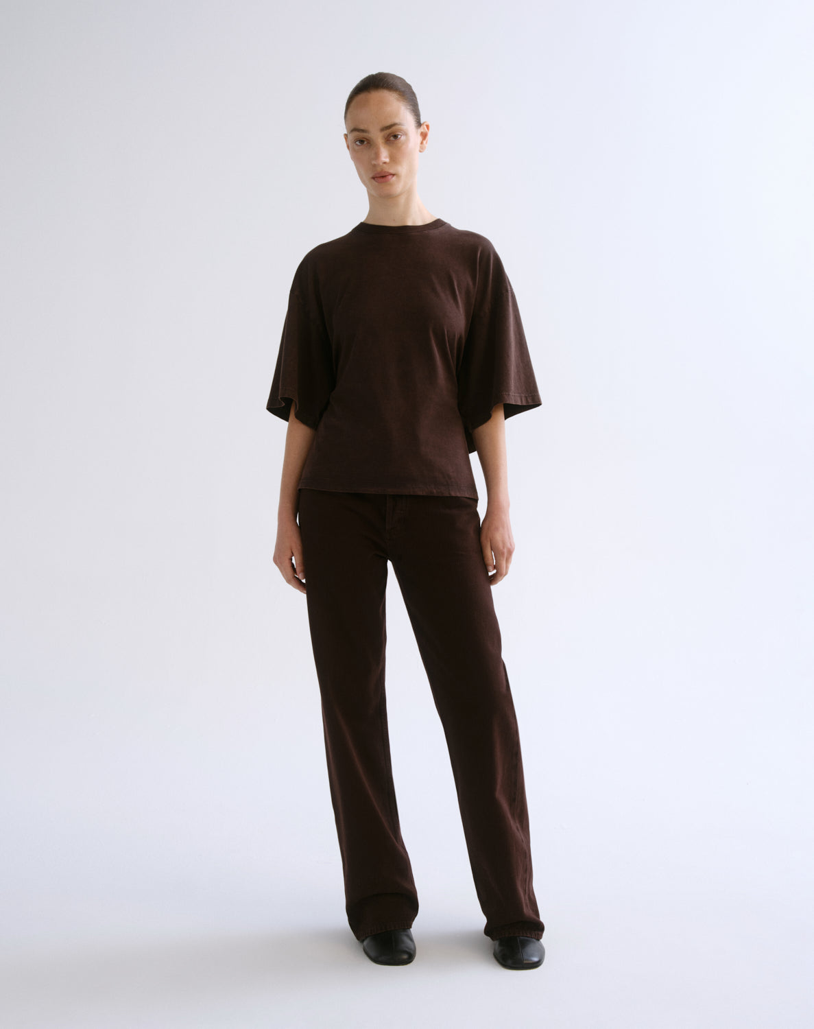 A model stands against a light gray studio background wearing a matching dark brown short-sleeve top and wide-leg trousers set.