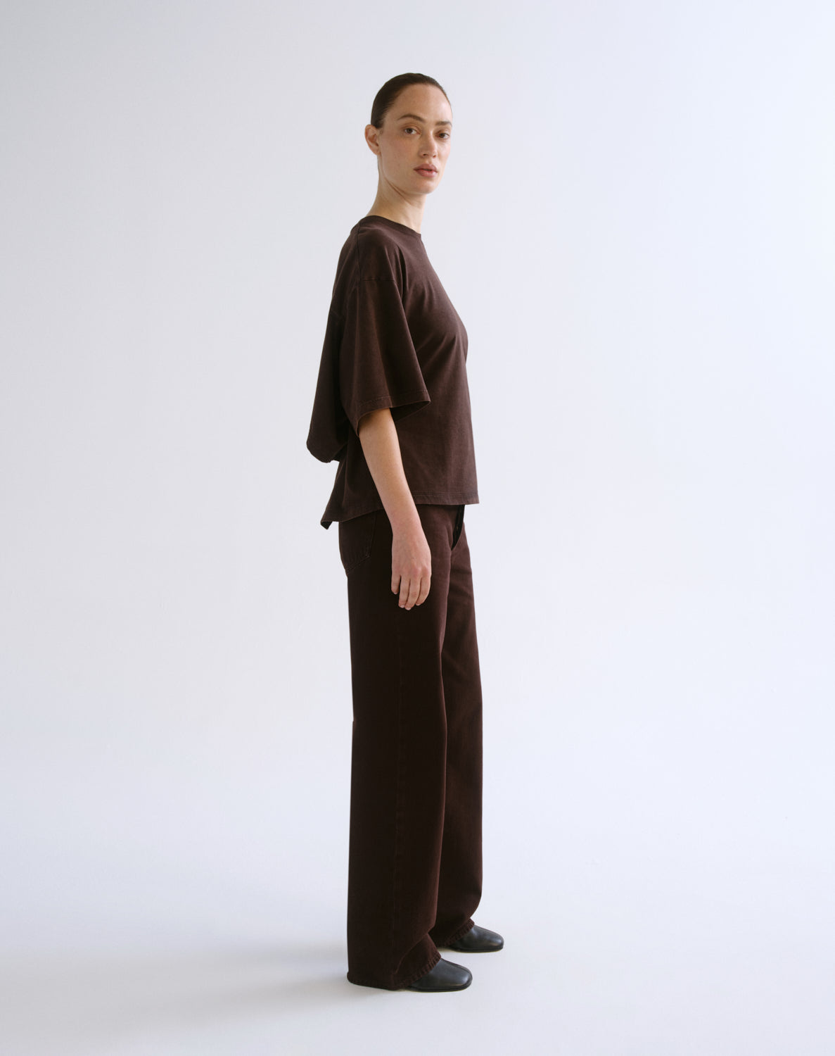 A model stands in profile wearing a dark brown oversized short-sleeve top and matching dark brown wide-leg pants against a plain light gray studio background.