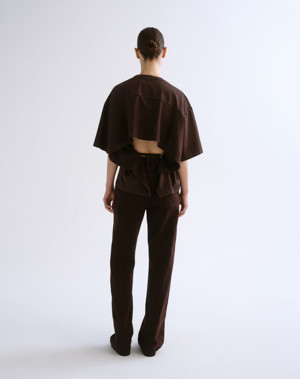 A model viewed from behind wearing a dark brown cropped short-sleeve top with a draped, layered back design and matching dark brown wide-leg trousers, set against a plain light gray studio background.
