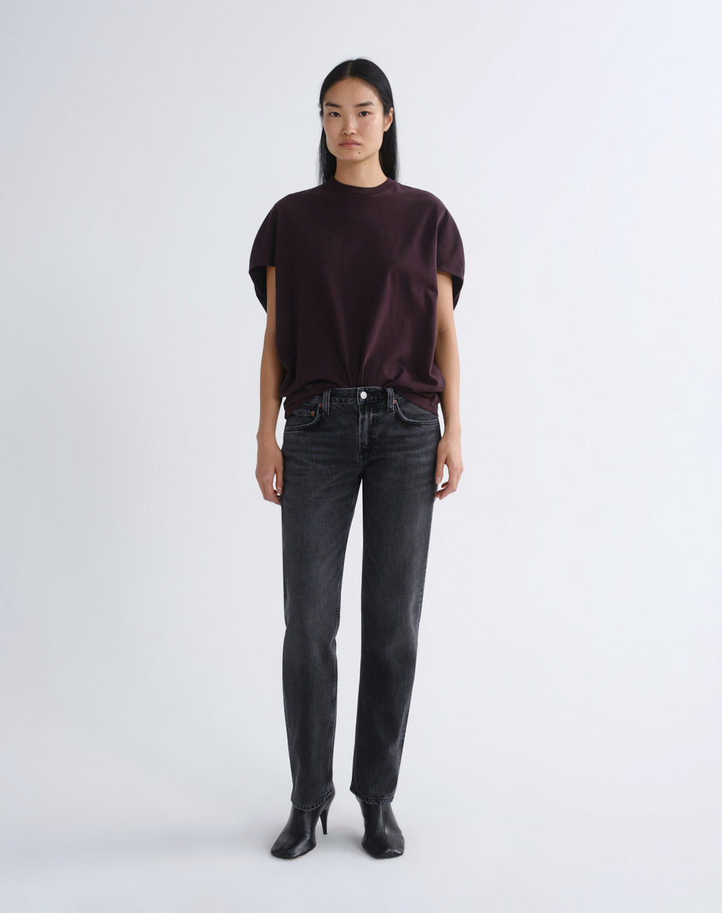 A model stands against a light gray studio background wearing a dark plum short-sleeve t-shirt, dark gray washed jeans, and black ankle boots.