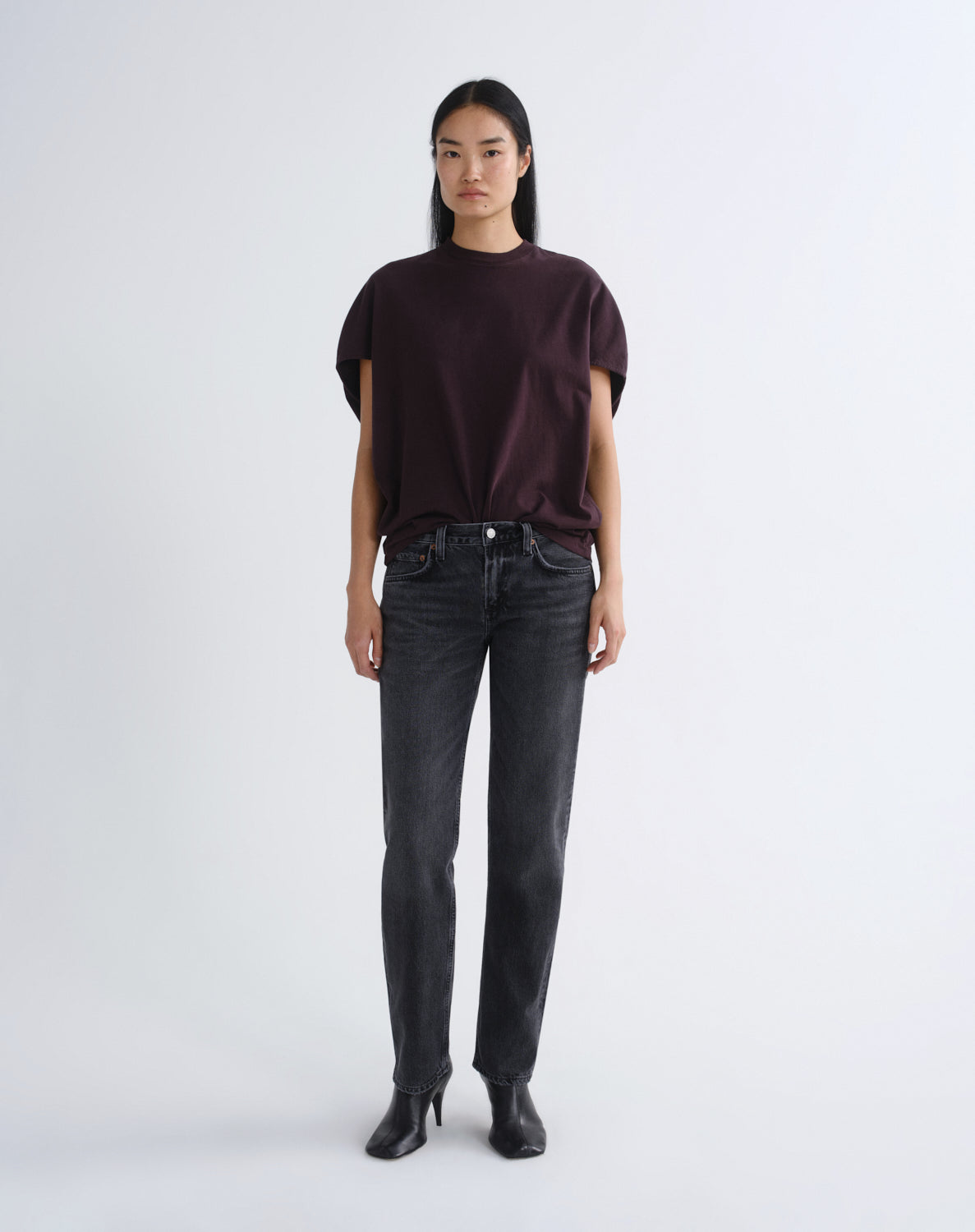 A model stands against a light gray studio background wearing a dark plum short-sleeve t-shirt, dark gray washed jeans, and black ankle boots.