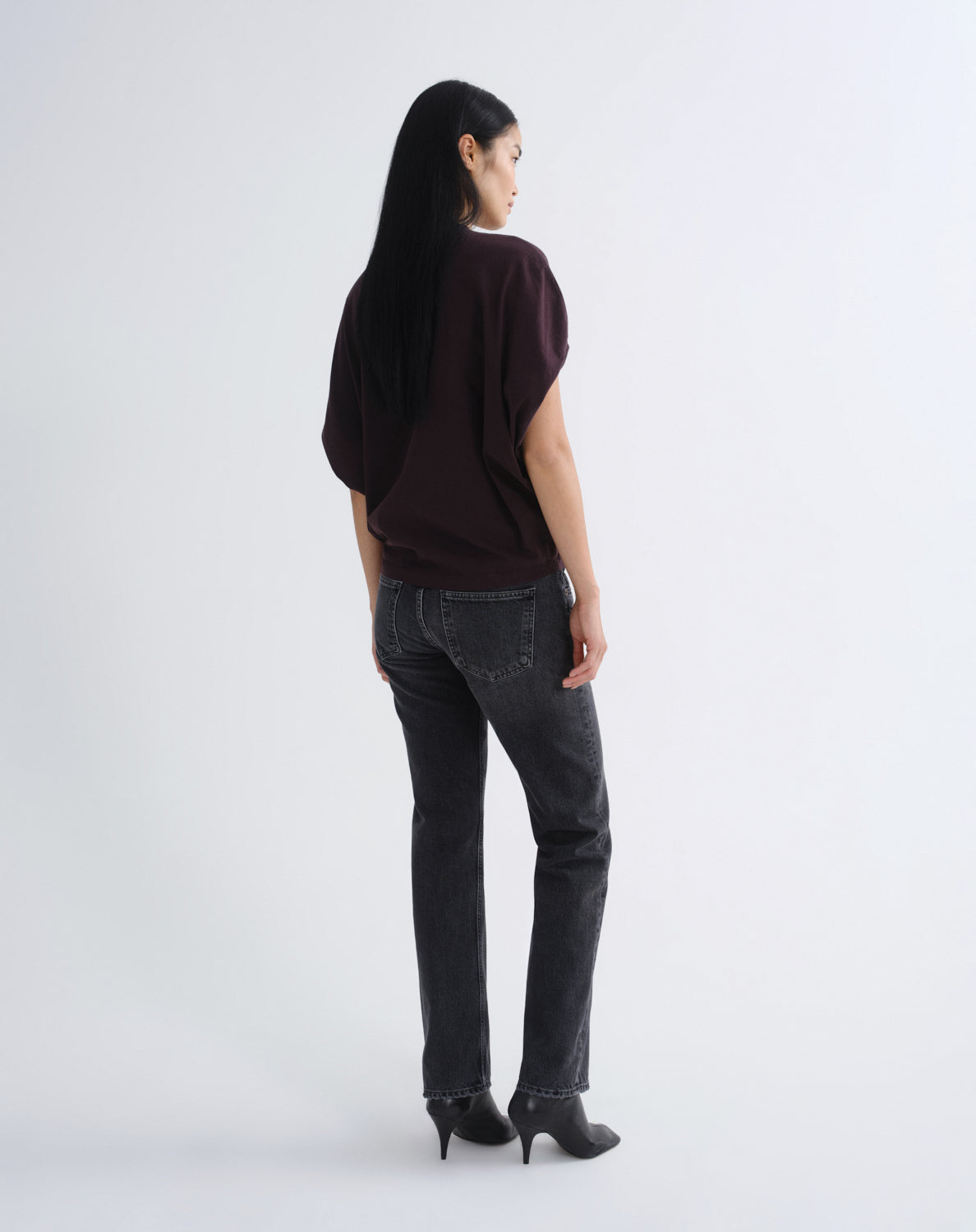 A woman with long black hair is shown from the back wearing a dark plum-purple short-sleeve top, dark gray straight-leg jeans, and black high-heeled boots against a plain light gray studio background.