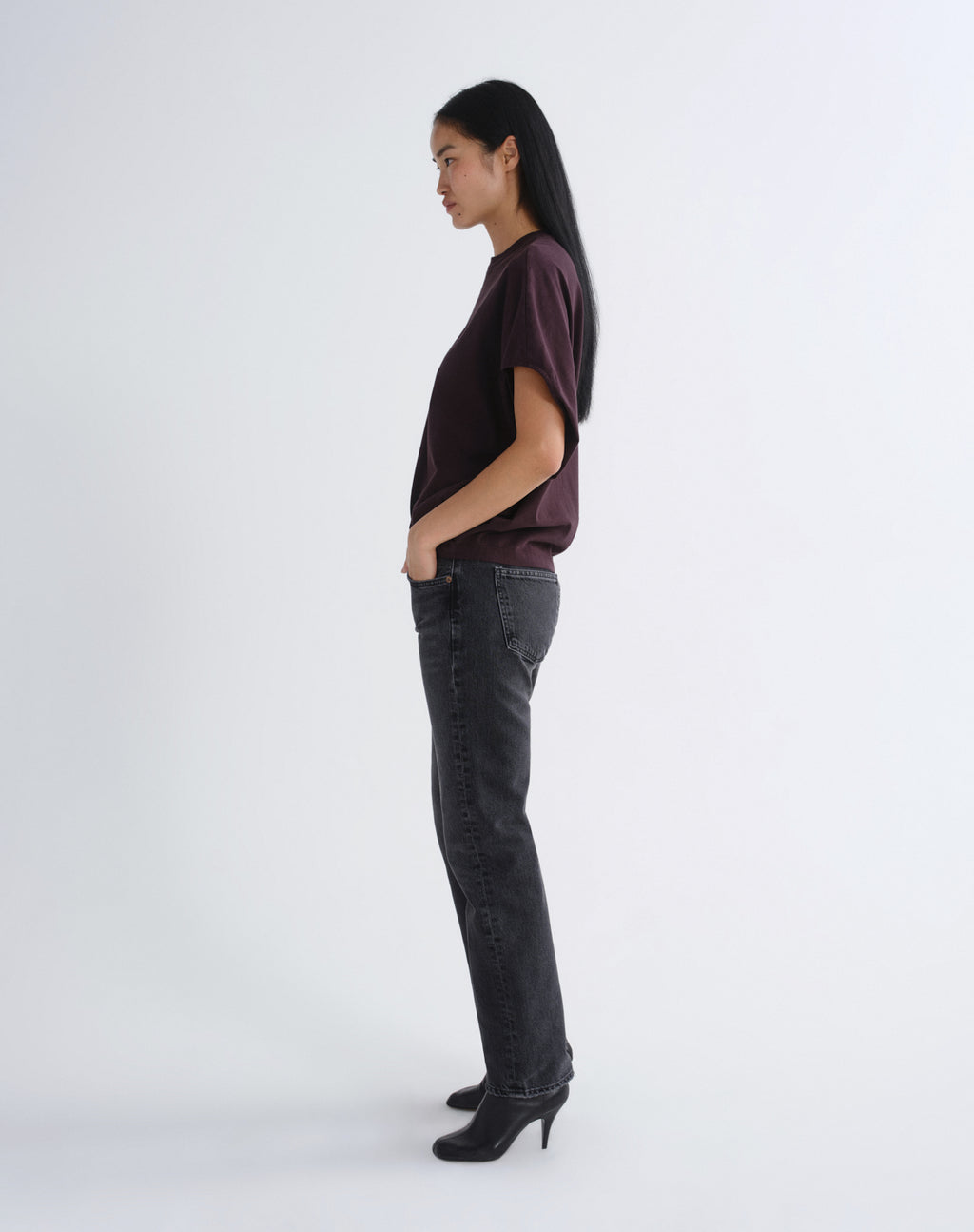 A woman stands in profile wearing a dark plum short-sleeve top, dark gray straight-leg jeans, and black pointed-toe heels against a plain light gray studio background.