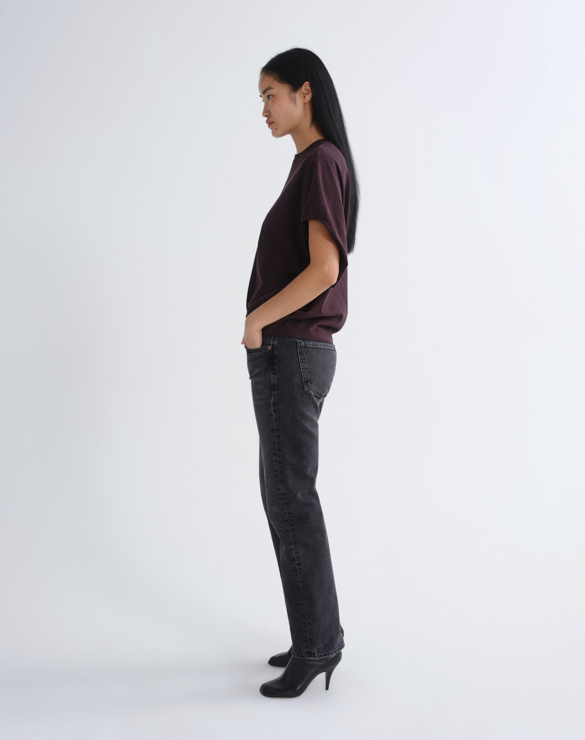 A woman stands in profile wearing a dark plum short-sleeve top, dark gray straight-leg jeans, and black pointed-toe heels against a plain light gray studio background.