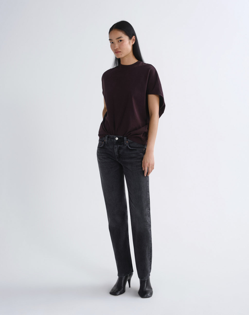 A model stands against a light gray studio background wearing a dark plum short-sleeve top, dark gray slim-fit jeans, and black ankle boots.