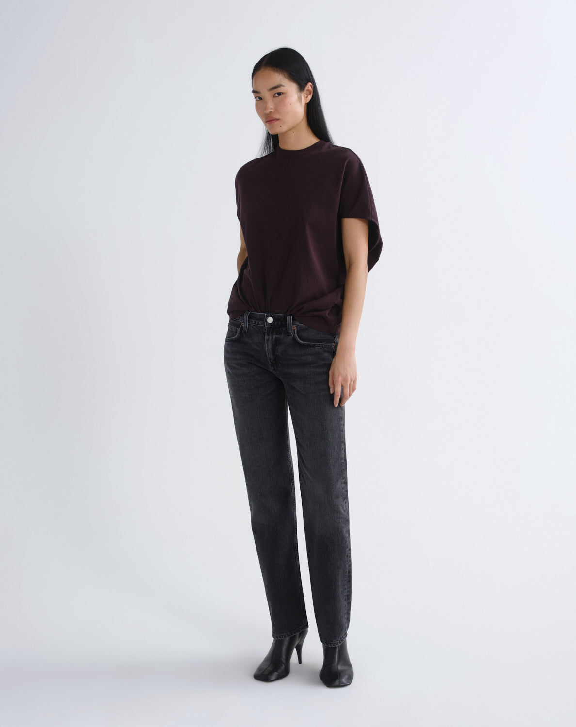 A model stands against a light gray studio background wearing a dark plum short-sleeve top, dark gray slim-fit jeans, and black ankle boots.