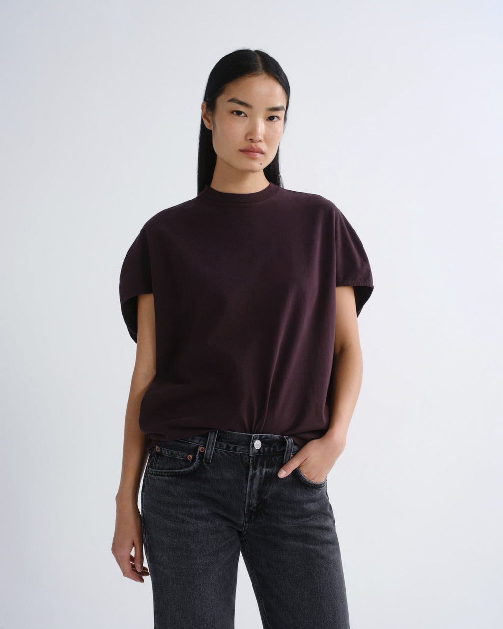 A model wearing a dark plum short-sleeve mock neck t-shirt and dark gray jeans, standing against a light gray studio background.