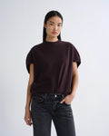A model wearing a dark plum short-sleeve mock neck t-shirt and dark gray jeans, standing against a light gray studio background.