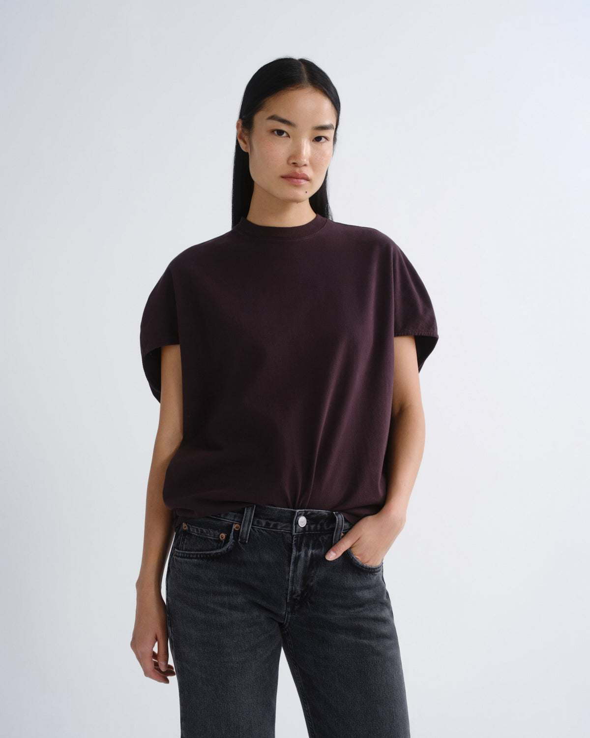 A model wearing a dark plum short-sleeve mock neck t-shirt and dark gray jeans, standing against a light gray studio background.