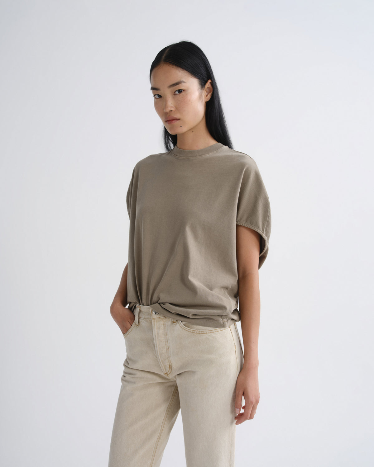 A model wearing a short-sleeve, crew-neck t-shirt in a muted taupe-brown color, paired with light beige jeans, against a plain light gray studio background.