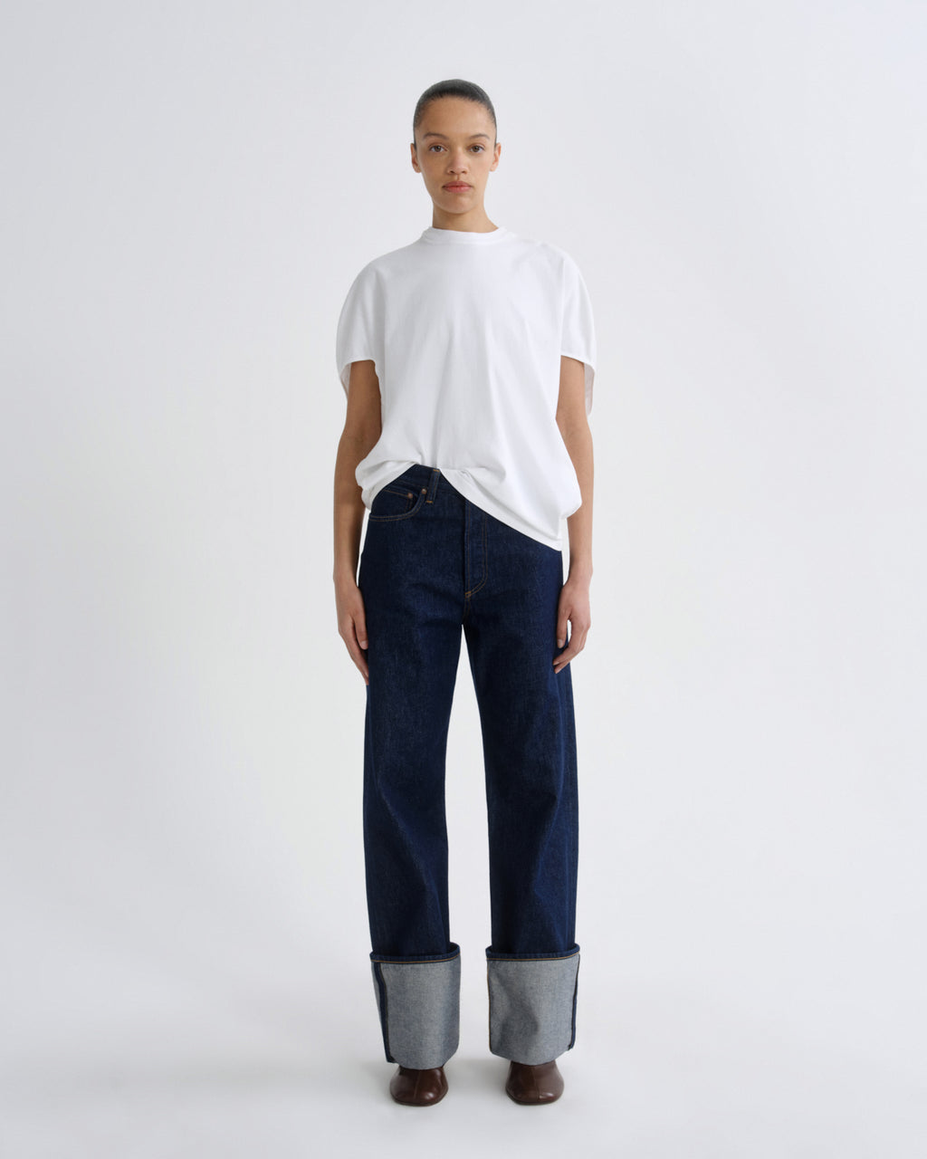 A model stands against a light gray studio background wearing a white short-sleeve t-shirt, dark blue straight-leg jeans with rolled cuffs showing a gray inner lining, and brown shoes.