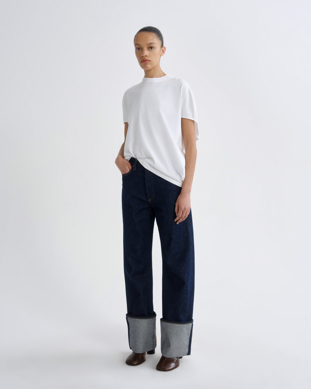 A model stands against a plain light gray studio background wearing a white short-sleeve crewneck t-shirt, dark blue wide-leg jeans with turned-up gray cuffs, and brown shoes.