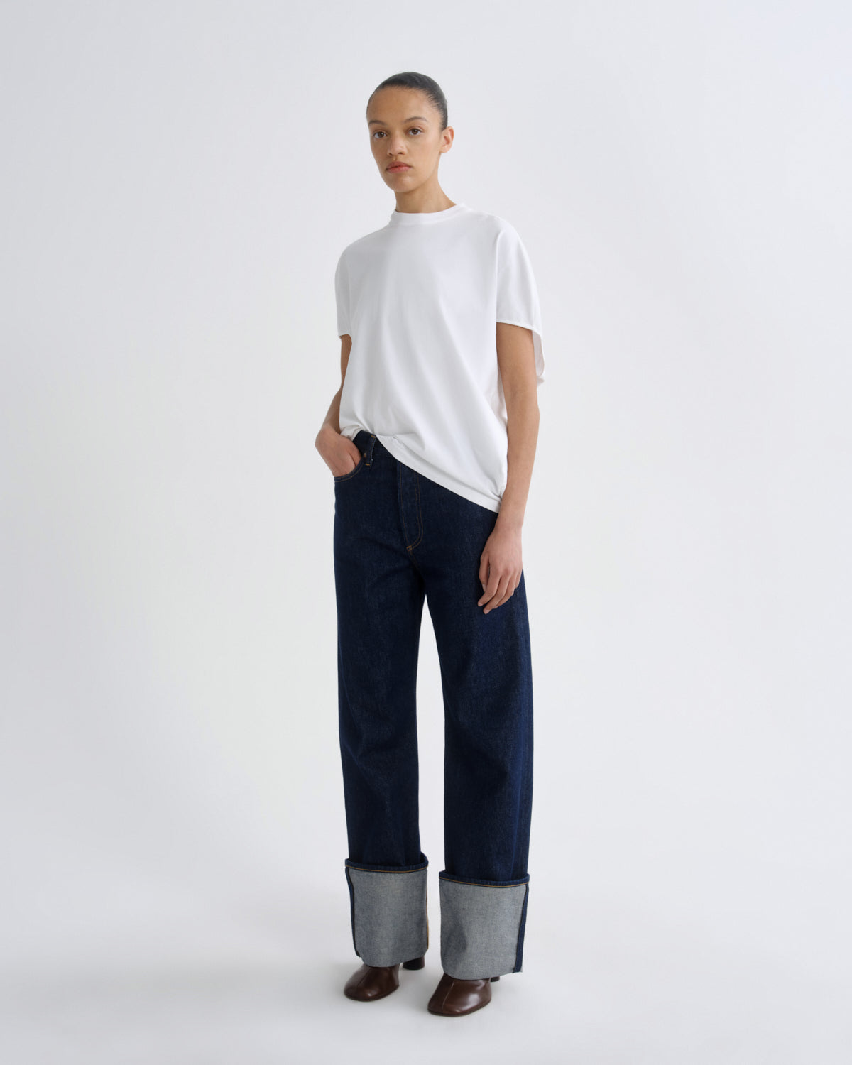 A model stands against a plain light gray studio background wearing a white short-sleeve crewneck t-shirt, dark blue wide-leg jeans with turned-up gray cuffs, and brown shoes.