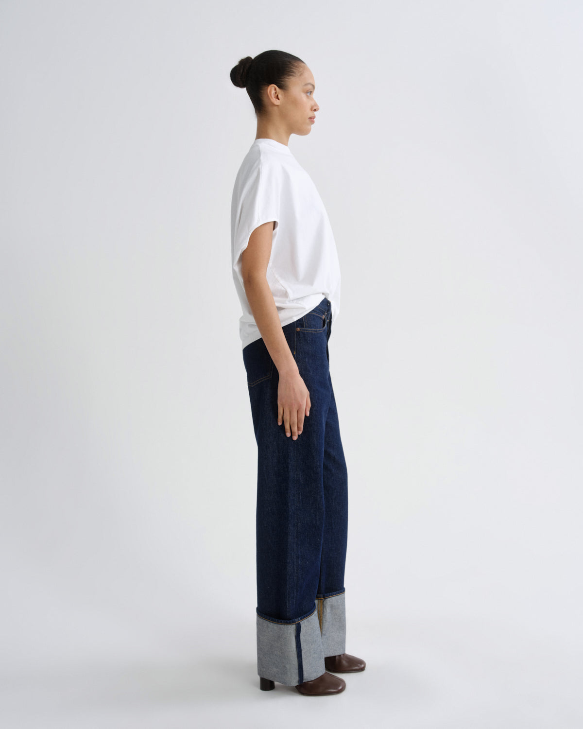 A model shown in profile wearing a white short-sleeve t-shirt, dark blue wide-leg jeans with cuffed hems, and brown shoes, set against a plain light gray studio background.