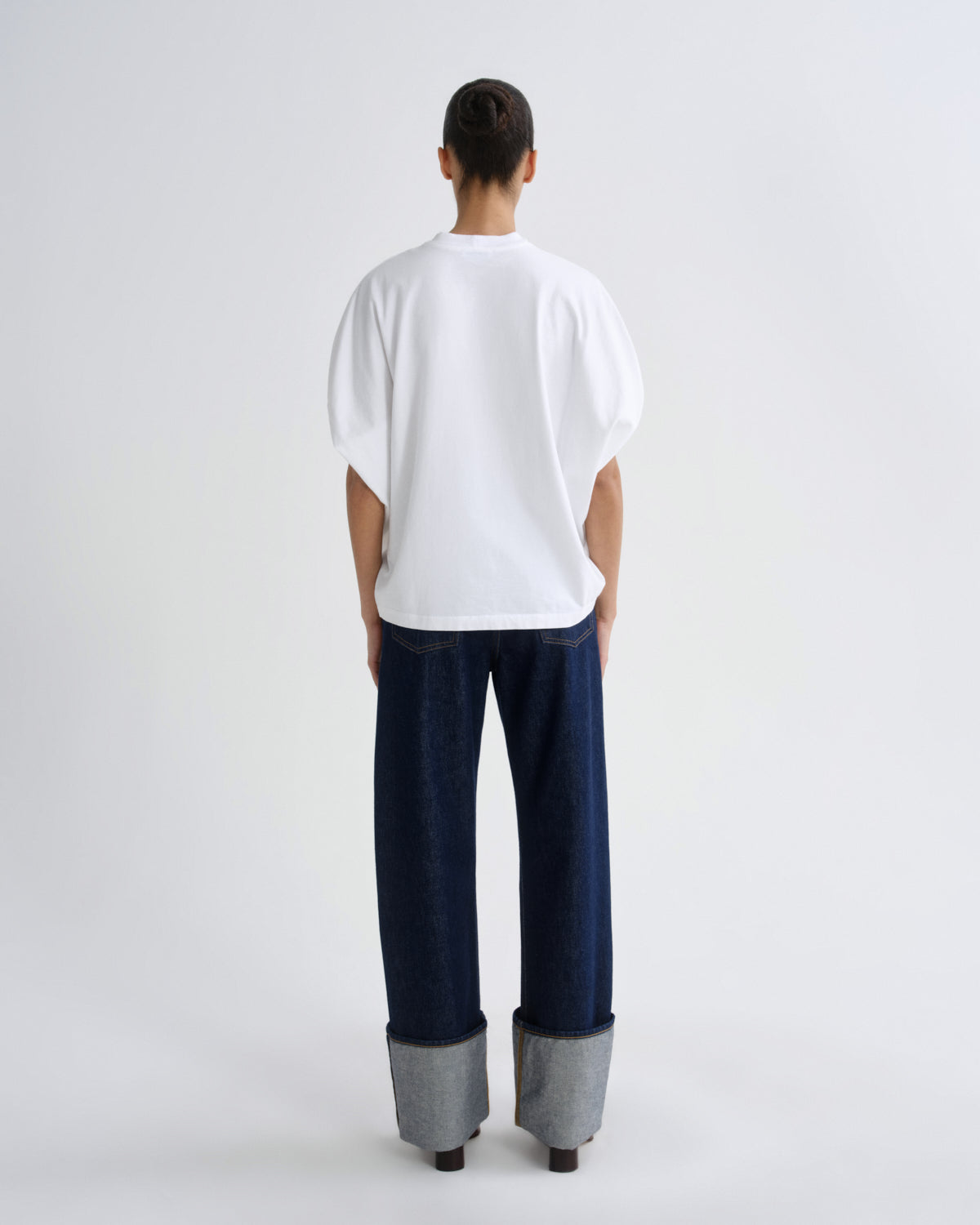 A model shown from the back wearing a white oversized short-sleeve t-shirt, dark blue wide-leg jeans with cuffed gray denim detailing, and black shoes, set against a plain light gray studio background.
