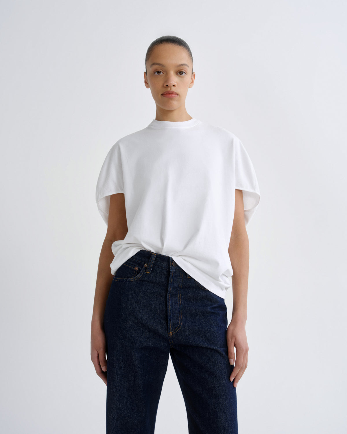 A model wearing a white short-sleeve crewneck t-shirt tucked into dark blue jeans, standing against a light gray studio background.