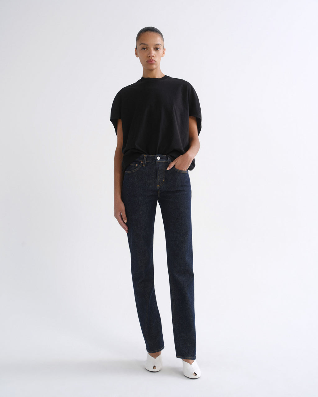 A model stands against a plain white studio background wearing a black short-sleeve t-shirt, dark blue straight-leg jeans, and white pointed shoes.