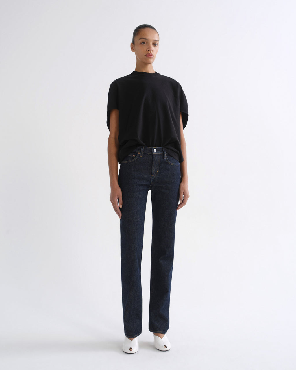 A model stands against a plain white studio background wearing a black short-sleeve t-shirt, dark blue straight-leg jeans, and white pointed shoes.