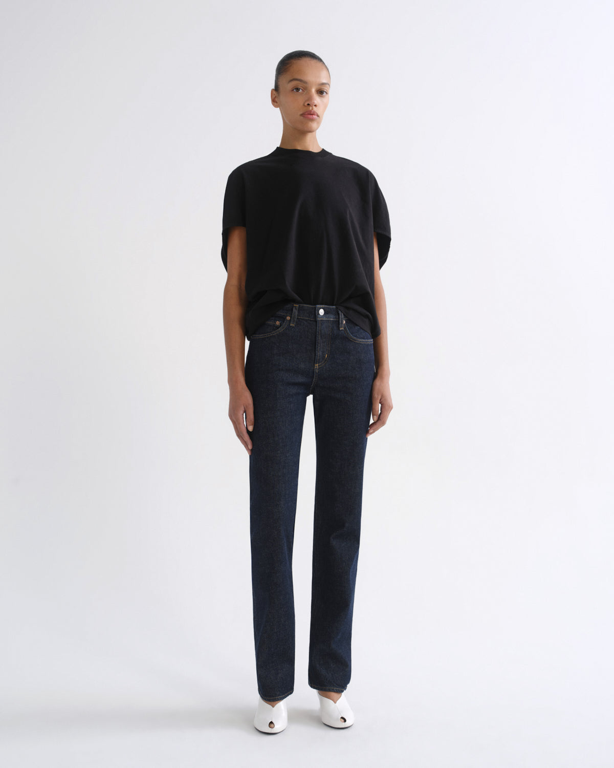 A model stands against a plain white studio background wearing a black short-sleeve t-shirt, dark blue straight-leg jeans, and white pointed shoes.