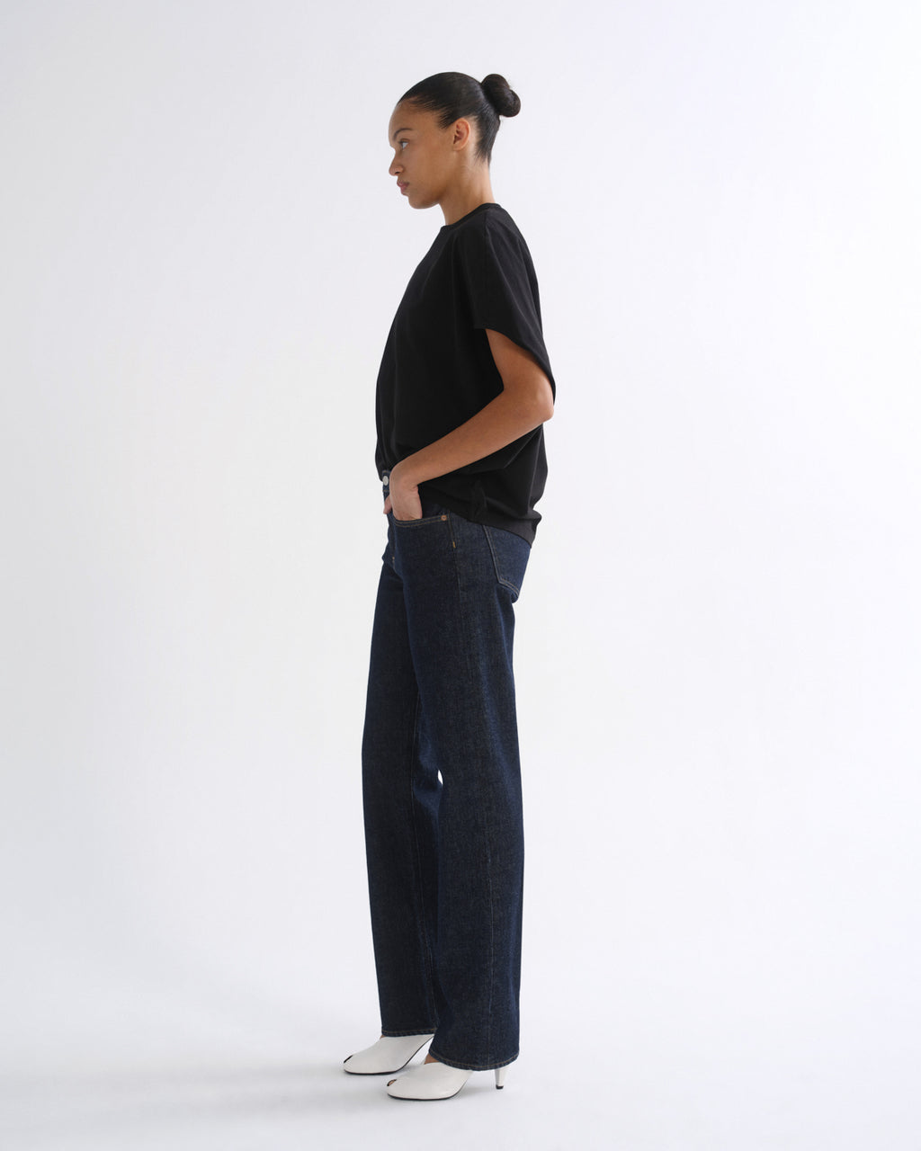 A model shown in profile wearing a black short-sleeve t-shirt, dark blue wide-leg denim jeans, and white pointed-toe heeled shoes, standing against a plain white studio background.