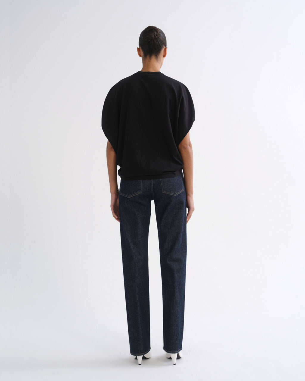 A model viewed from the back wearing a black short-sleeve top and dark blue straight-leg jeans, standing against a plain white studio background.