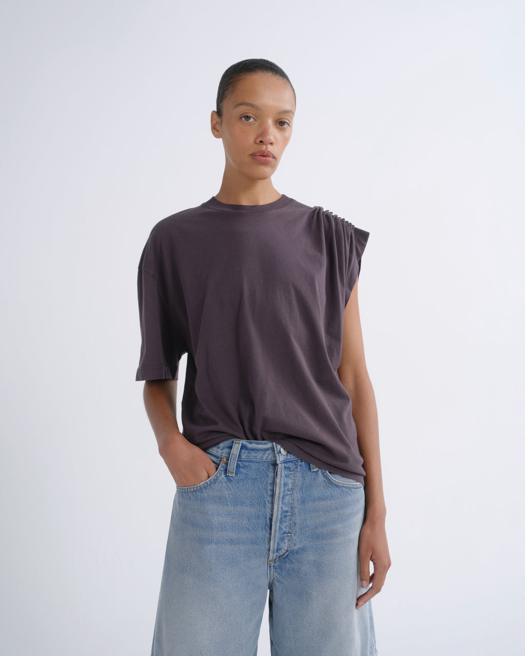 A model wearing a dark plum-purple oversized t-shirt with a unique asymmetrical shoulder design and light-wash blue jeans, standing against a light gray studio background.