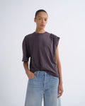 A model wearing a dark plum-purple oversized t-shirt with a unique asymmetrical shoulder design and light-wash blue jeans, standing against a light gray studio background.