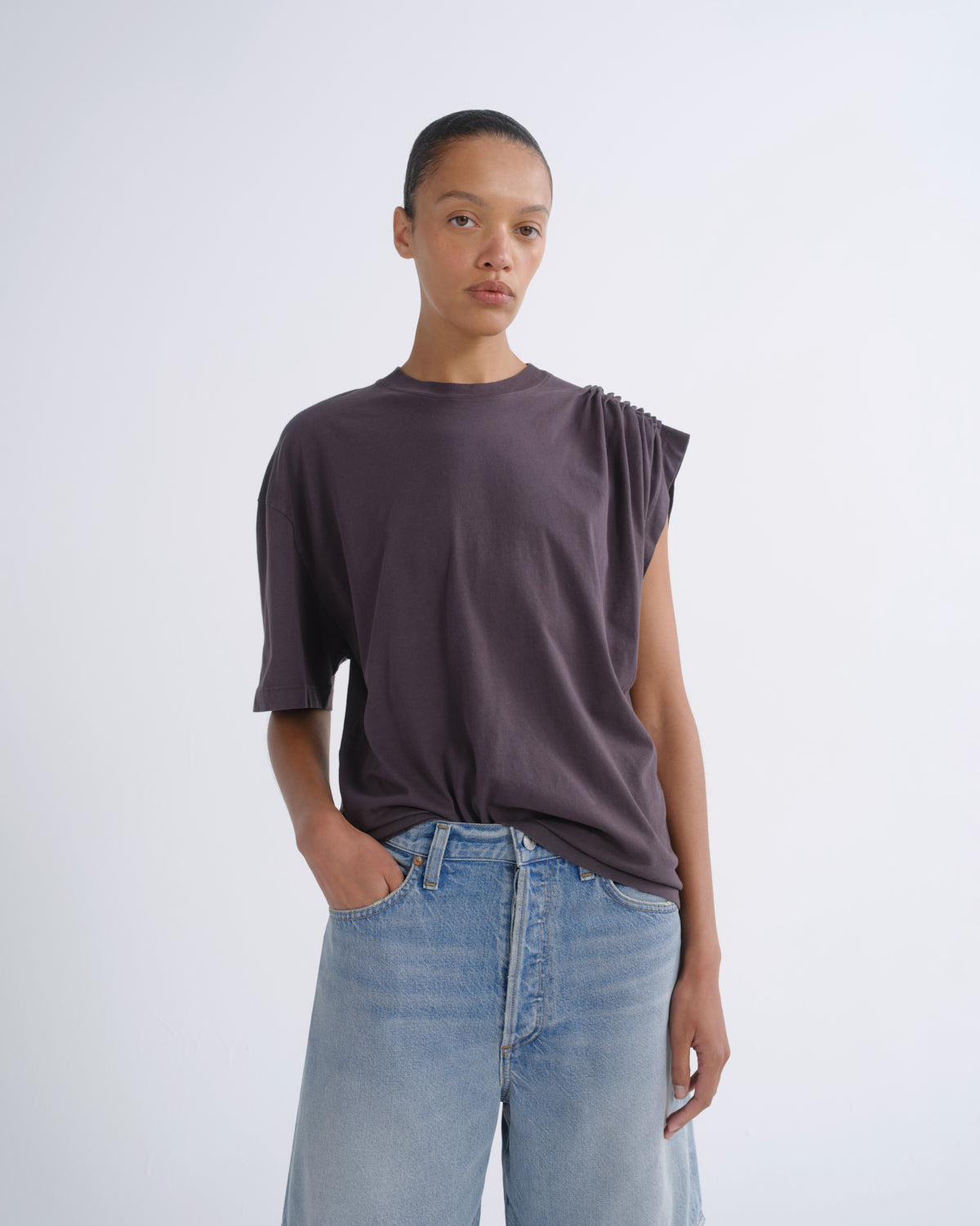 A model wearing a dark plum-purple oversized t-shirt with a unique asymmetrical shoulder design and light-wash blue jeans, standing against a light gray studio background.