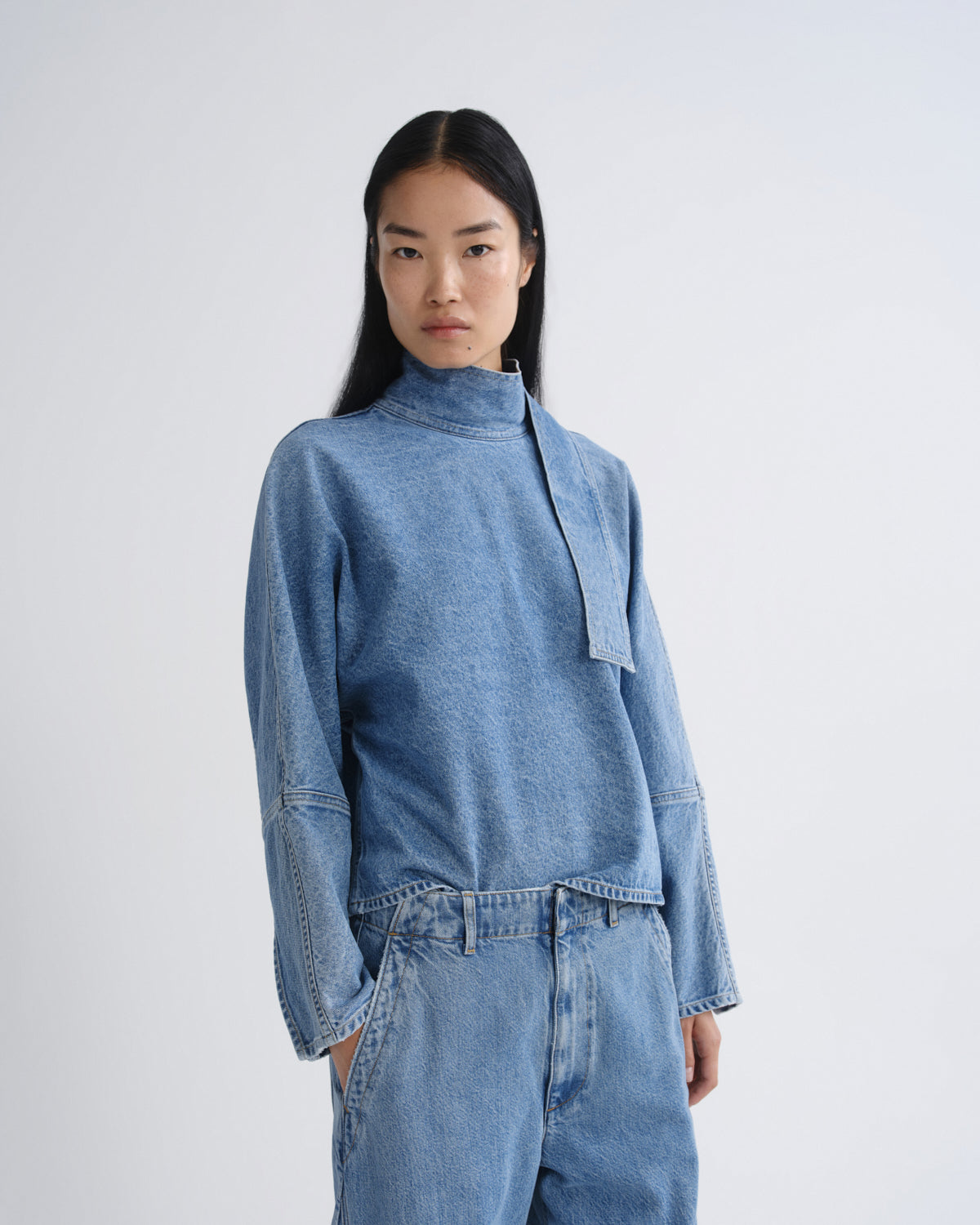 A model wearing a light blue denim long-sleeve top with a high mock neck and matching wide-leg denim jeans, photographed against a solid light gray background.