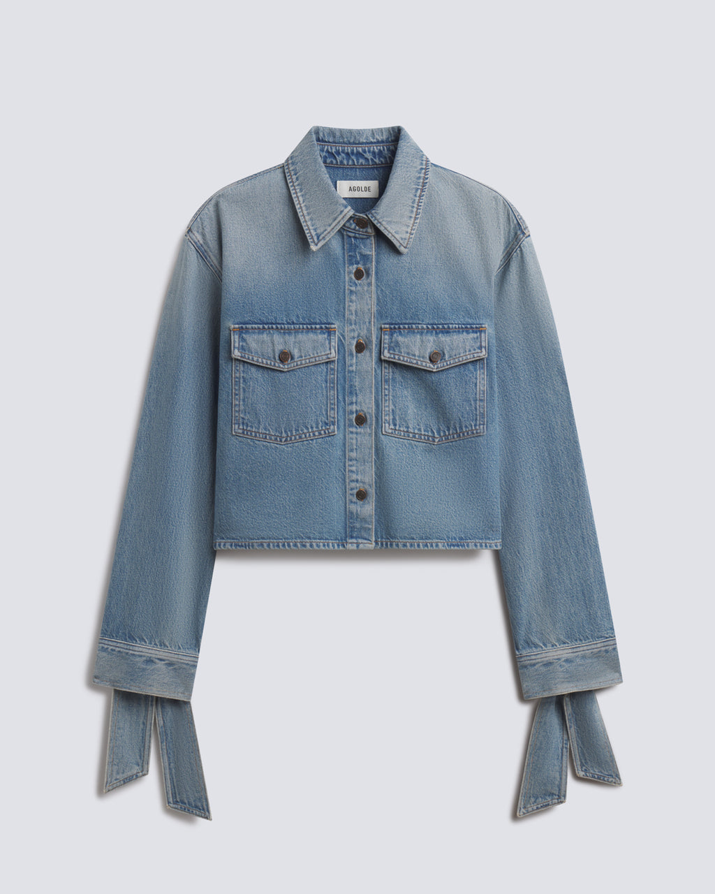 A cropped light blue denim jacket with long sleeves, button-front closure, two flap chest pockets, and matching fabric tie details at the cuffs, shown on a light gray background.