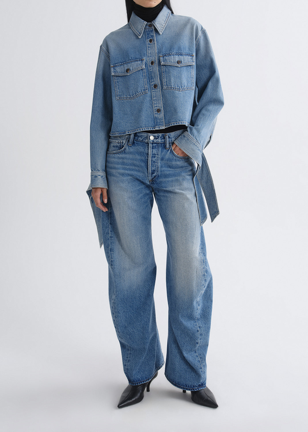 A model wearing a light blue cropped denim shirt-jacket with matching wide-leg jeans, styled with a black turtleneck and black pointed-toe heels against a grey background.