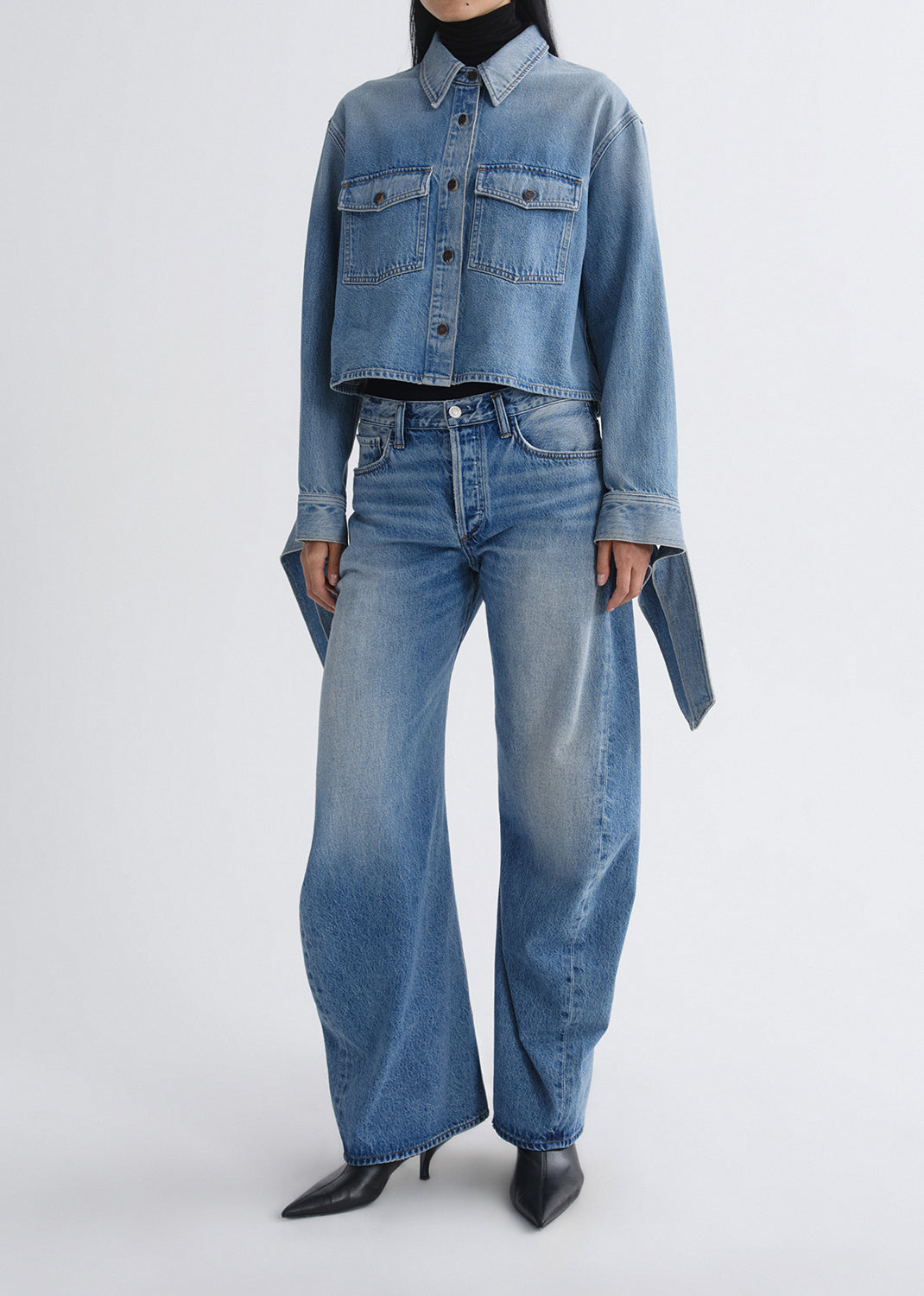 A model wearing a light blue cropped denim shirt-jacket with long sleeves and matching wide-leg denim jeans, styled with black pointed-toe heels against a grey background.