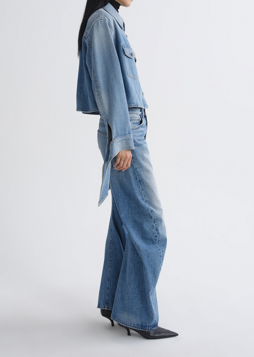A model shown from the side wearing a light blue denim jacket with a tie detail and matching wide-leg denim jeans, paired with black pointed-toe heels, against a grey background.