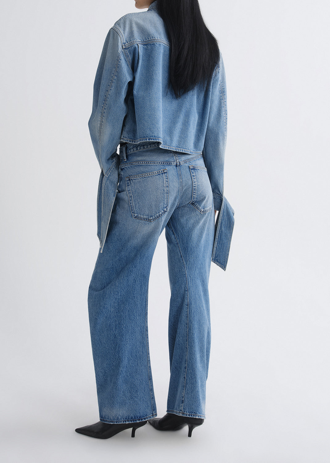 A model shown from the back wearing a light blue denim outfit, featuring a cropped denim jacket with long sleeves and matching wide-leg denim jeans, paired with black pointed-toe heels against a grey background.
