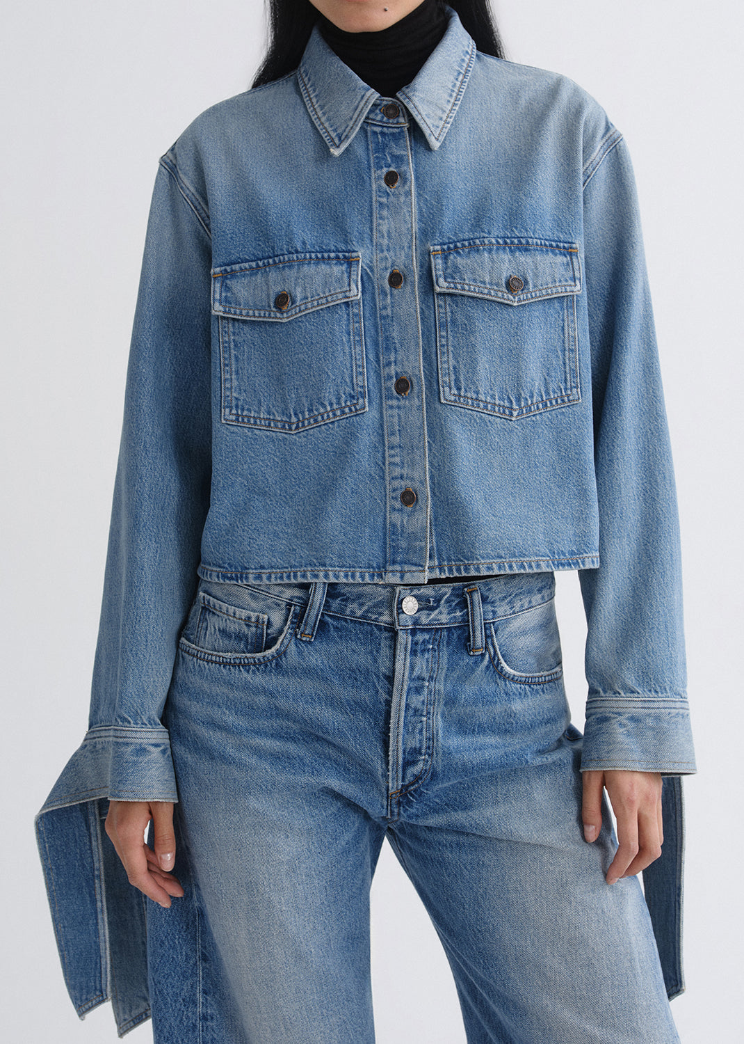 A model wearing a cropped light blue denim shirt jacket with front flap pockets, paired with matching blue jeans and a black turtleneck, shown against a grey background.