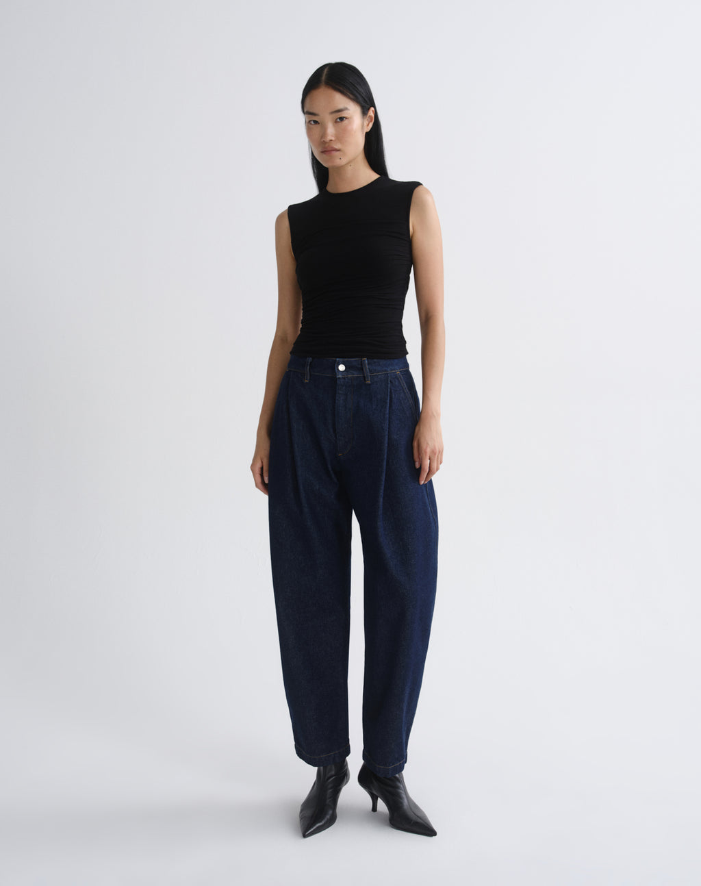 A model stands against a grey  background wearing a sleeveless black ribbed top, dark navy wide-leg jeans, and black pointed-toe heeled boots.
