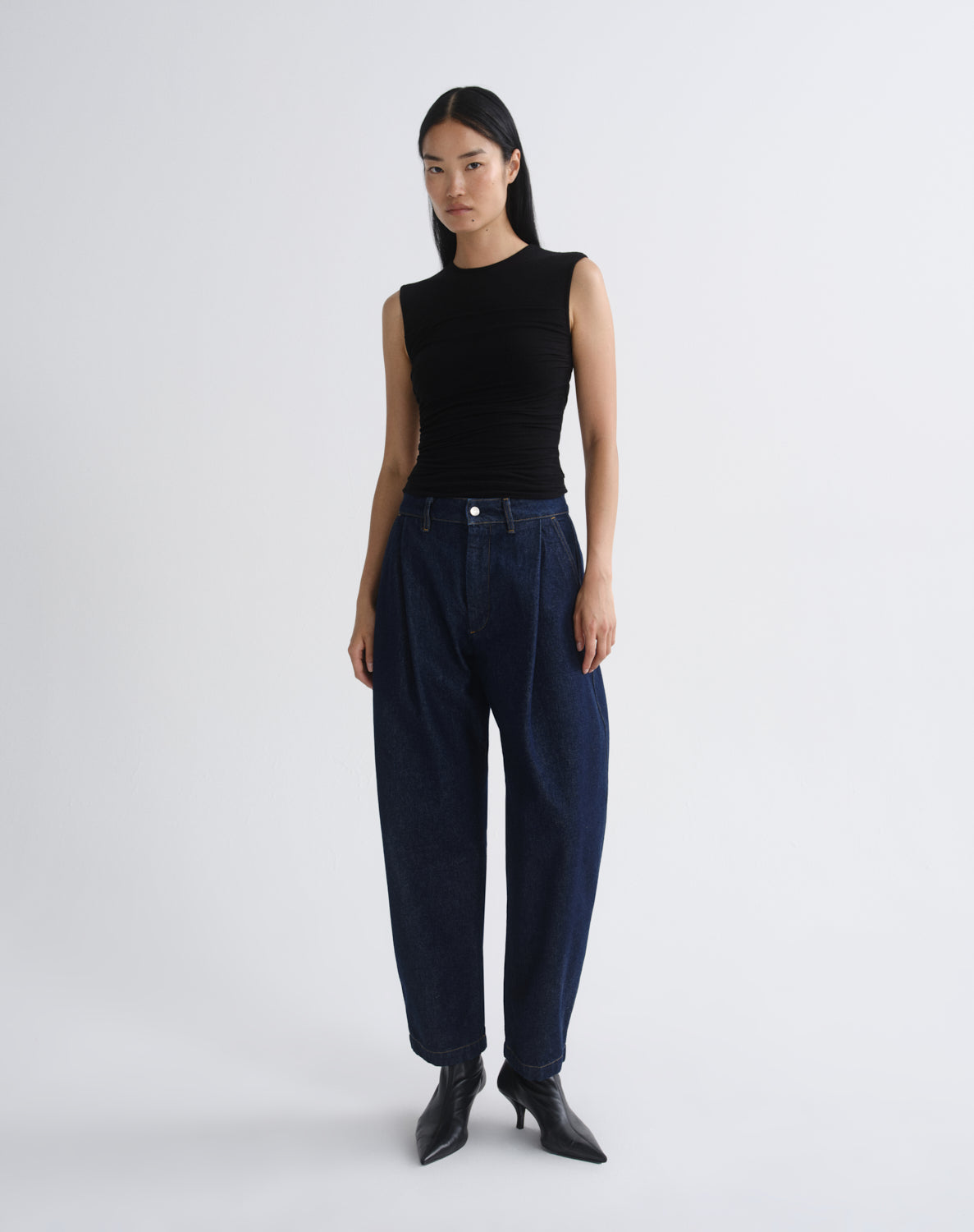 A model stands against a grey  background wearing a sleeveless black ribbed top, dark navy wide-leg jeans, and black pointed-toe heeled boots.