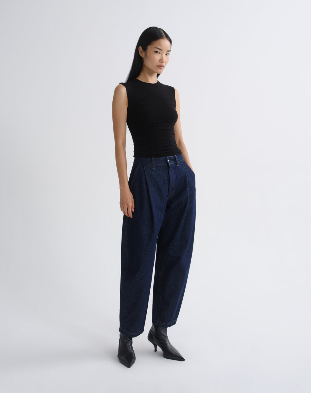 A model stands in profile wearing a sleeveless black top, dark navy wide-leg trousers, and black pointed ankle boots against a grey background.
