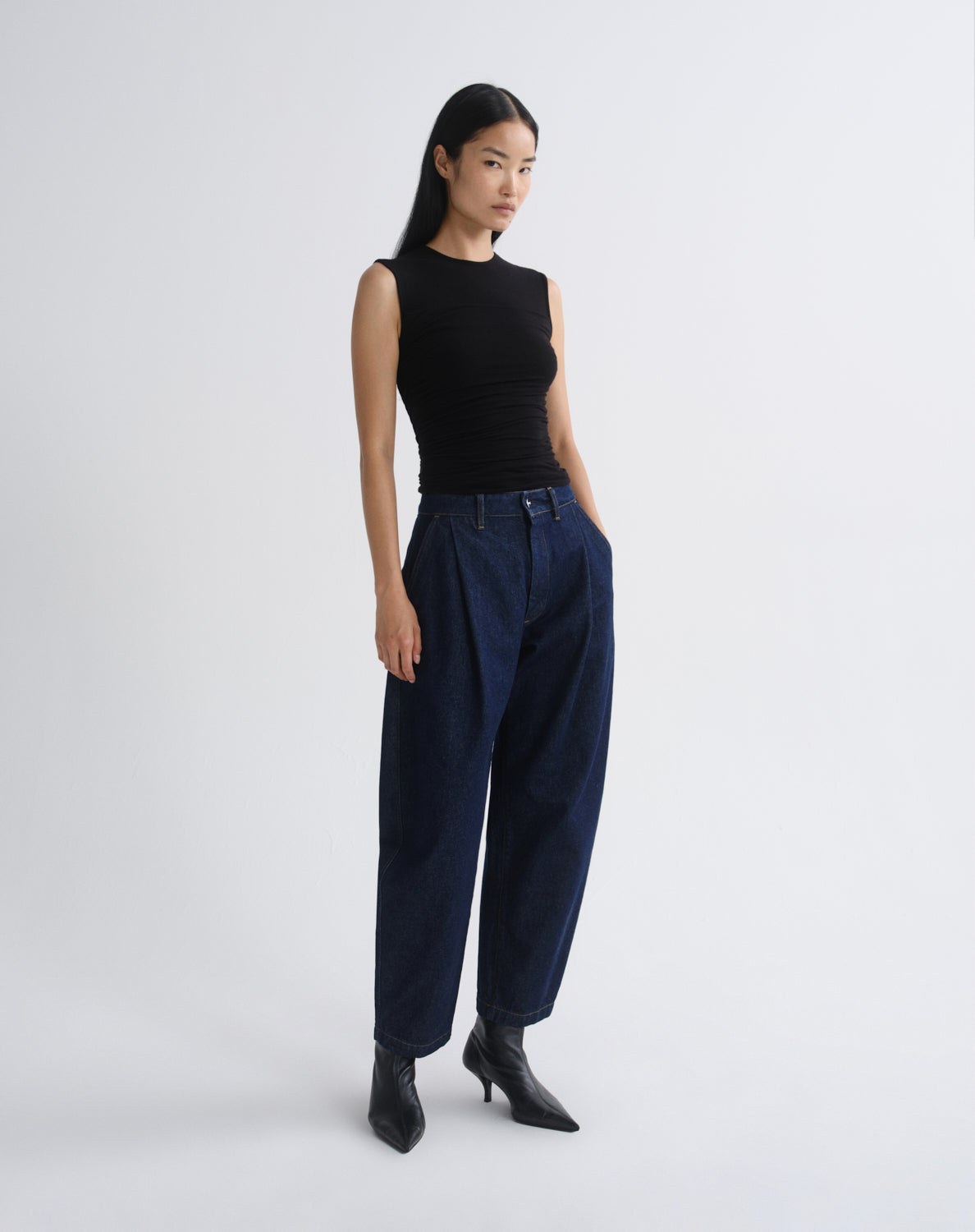 A model stands in profile wearing a sleeveless black top, dark navy wide-leg trousers, and black pointed ankle boots against a grey background.