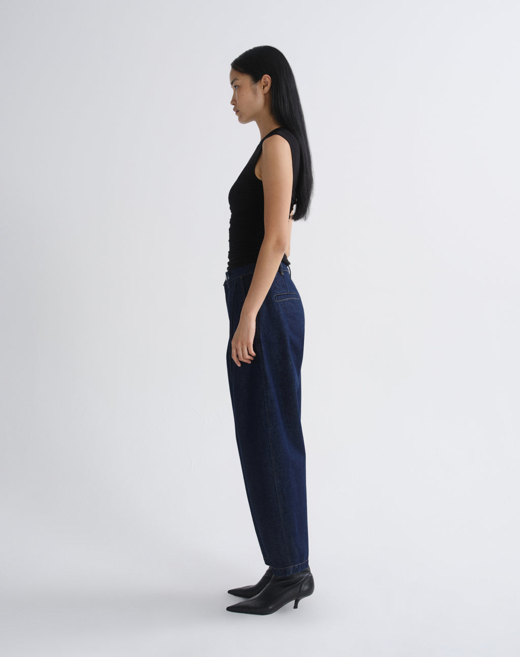A woman shown in profile wearing dark blue wide-leg jeans, a black sleeveless top, and black pointed-toe heeled boots against a grey background.
