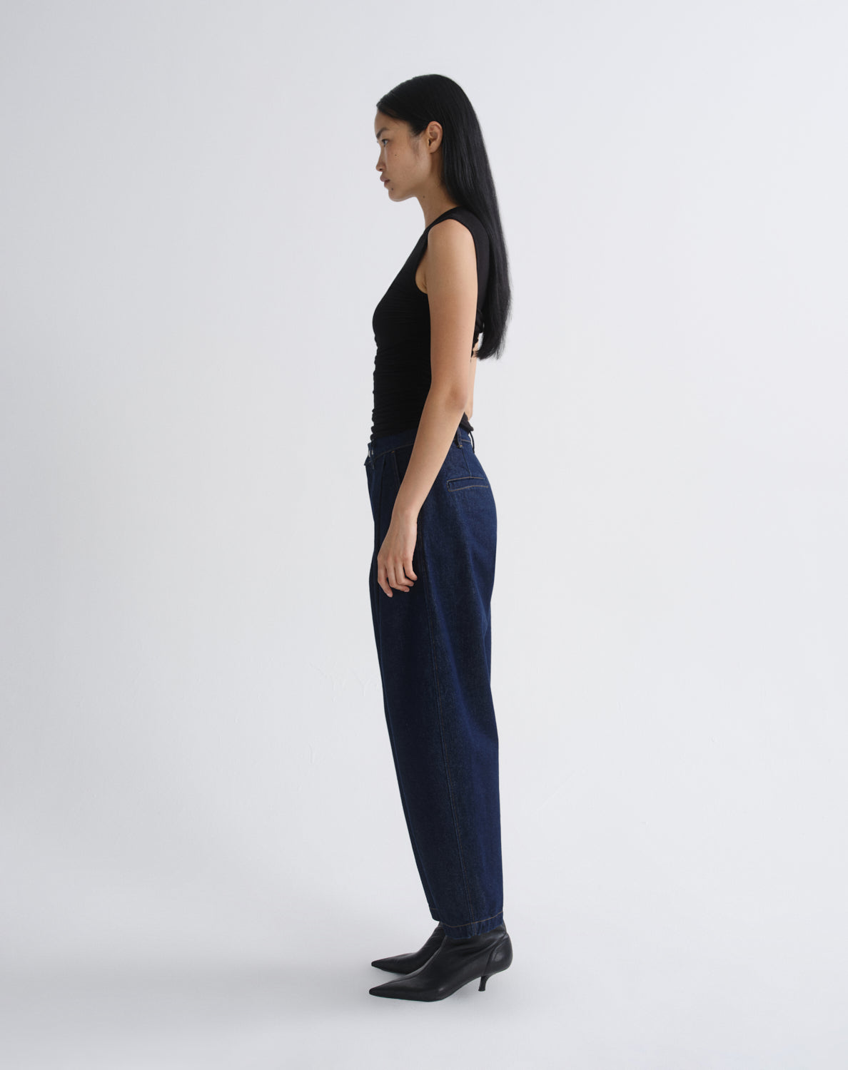 A woman shown in profile wearing dark blue wide-leg jeans, a black sleeveless top, and black pointed-toe heeled boots against a grey background.