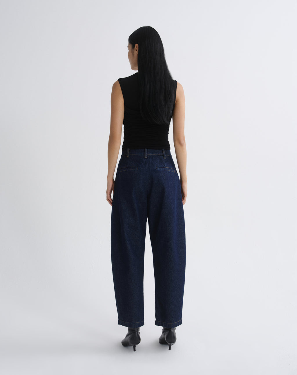 A woman viewed from behind wearing a black sleeveless top and dark blue wide-leg jeans with black pointed-toe heels, standing against  grey background.