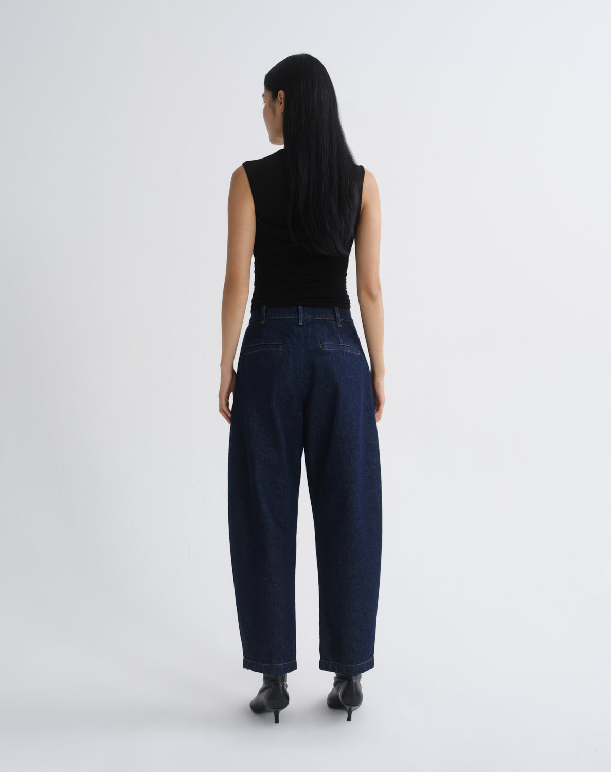 A woman viewed from behind wearing a black sleeveless top and dark blue wide-leg jeans with black pointed-toe heels, standing against  grey background.
