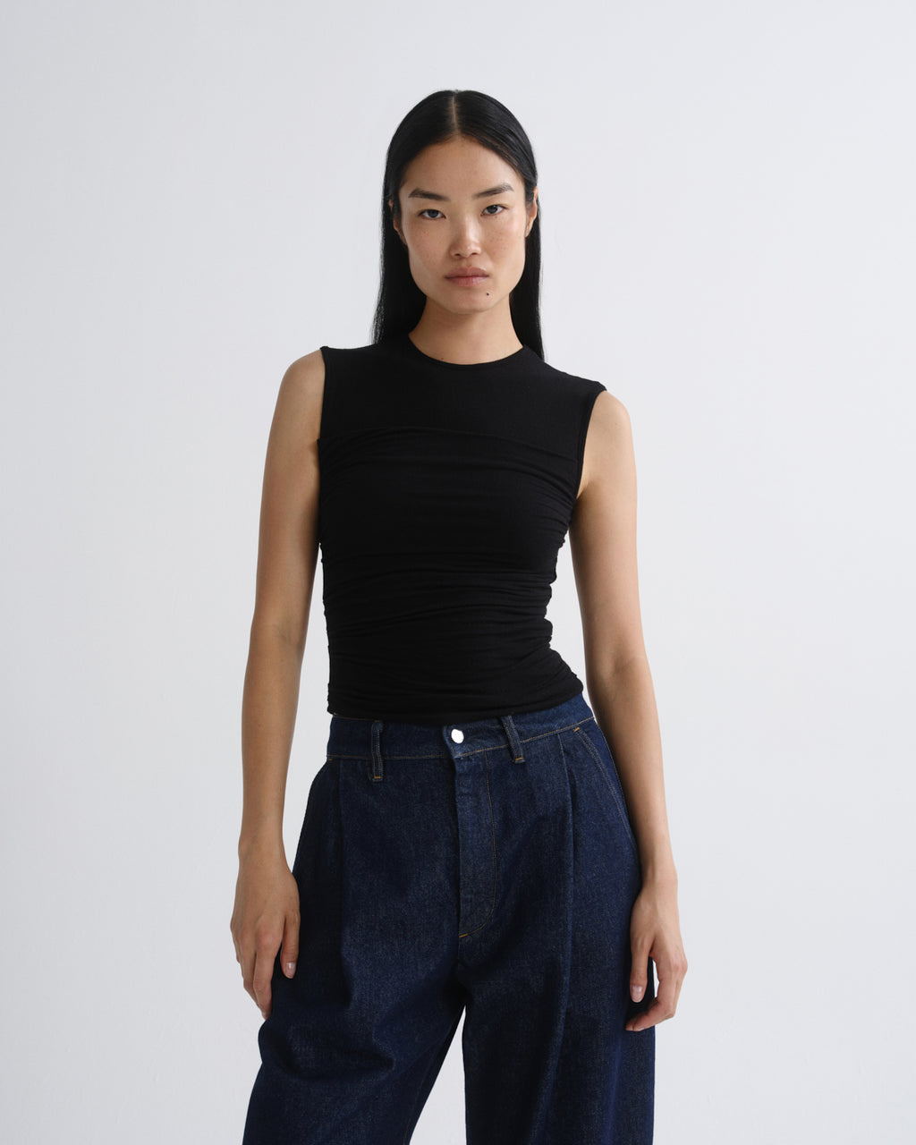 A model wearing a black sleeveless ribbed top and dark blue wide-leg jeans, standing against grey background.