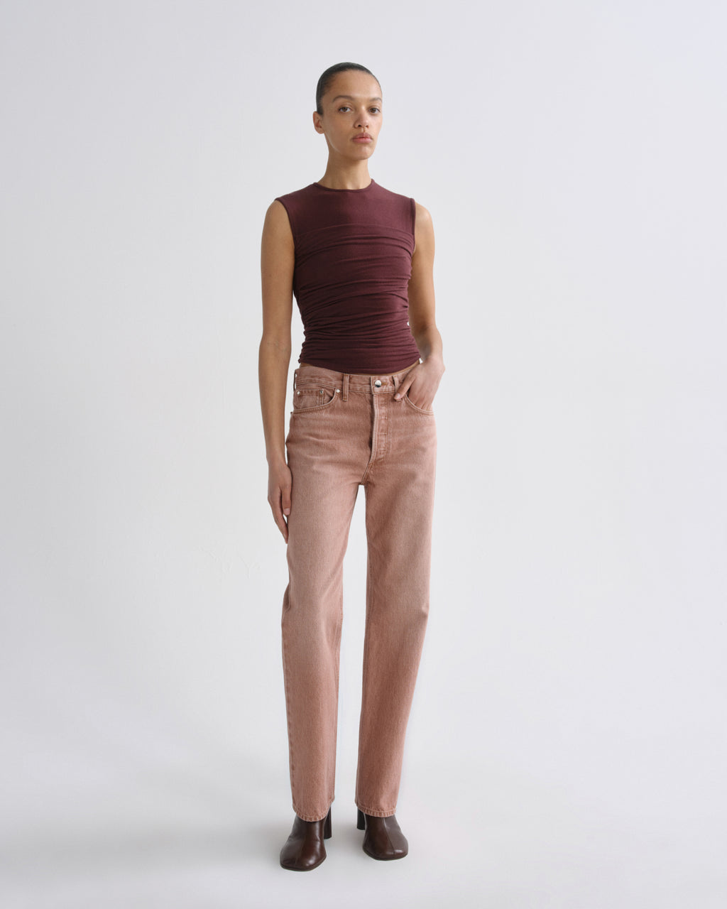 A model stands against a light gray studio background wearing a sleeveless, ruched burgundy top and straight-leg dusty rose jeans with dark brown ankle boots.