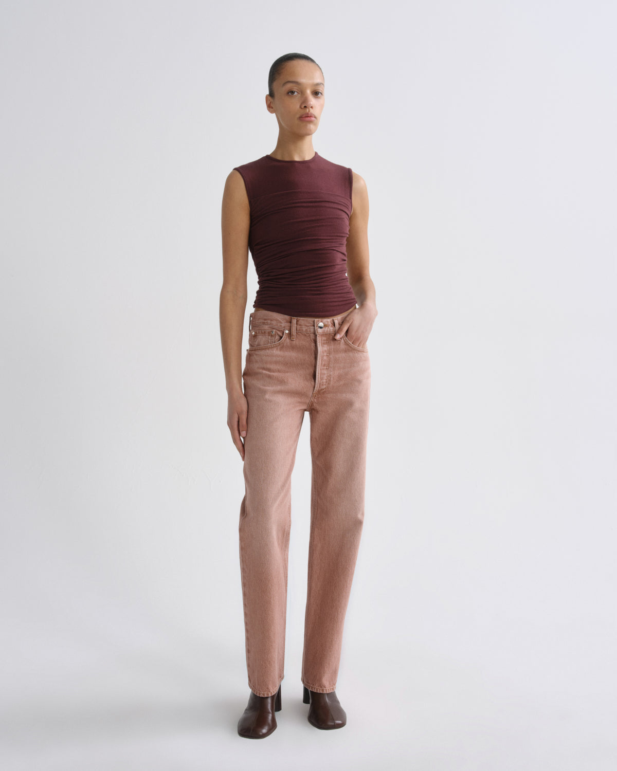 A model stands against a light gray studio background wearing a sleeveless, ruched burgundy top and straight-leg dusty rose jeans with dark brown ankle boots.